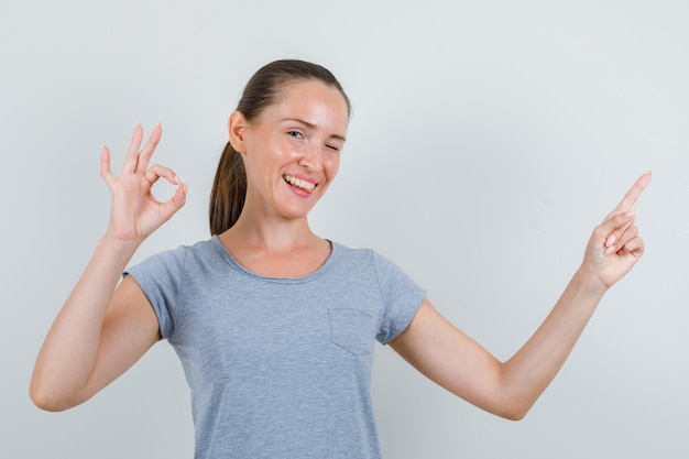 Junge Frau, die zur Seite mit OK-Zeichen in grauem T-Shirt, Brille zeigt und froh schaut. Vorderansicht.