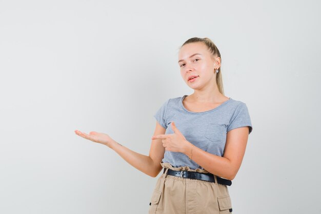 Junge Frau, die zur Seite zeigt, vorgibt, etwas im T-Shirt, in der Hose zu halten und selbstbewusst, Vorderansicht aussehend.