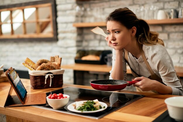 Junge Frau fühlt sich beim Kochen unzufrieden und schaut sich das Rezept im Internet an