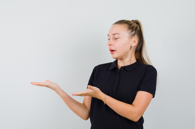 Junge Frau gestikuliert, als würde sie etwas in einem schwarzen T-Shirt präsentieren und attraktiv aussehen