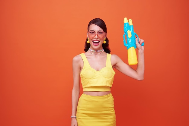 Junge Frau glückliche Schönheit mit Wasserpistole und während Songkran Festival Studioaufnahme isoliert auf orangefarbenem Hintergrund