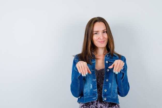 Junge Frau hält die Hände über der Brust als Handflächen in Jeansjacke, Kleid und sieht positiv aus. Vorderansicht.