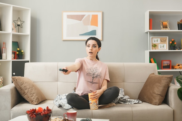 Junge Frau hält die TV-Fernbedienung zur Kamera auf dem Sofa hinter dem Couchtisch im Wohnzimmer