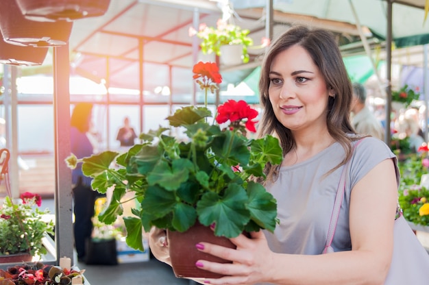 Junge Frau hält Pelargonie in Tontopf im Garten Zentrum. Gartenarbeit, Pflanzung - Frau mit Pelargonie blüht