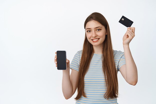 Junge Frau hebt die Hand mit Kreditkarte und lächelt zufrieden, zeigt Smartphone-Bildschirm, Handy-App-Schnittstelle für Banking auf Weiß.