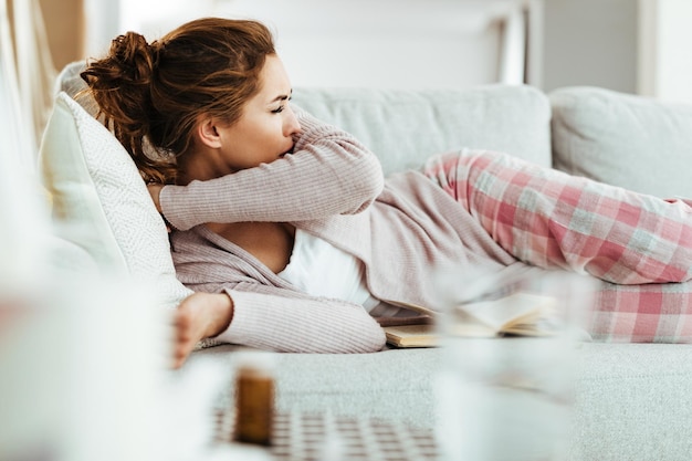 Junge Frau hustet in den Ellbogen, während sie sich auf dem Sofa im Wohnzimmer hinlegt