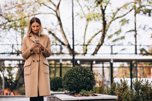 Junge Frau im beige Mantel unter Verwendung des Telefons außerhalb der Straße