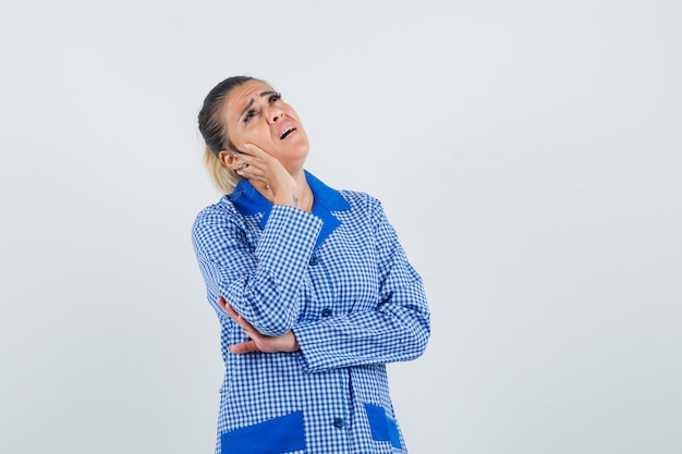 Junge Frau im blauen Gingham-Pyjamahemd, das Hand auf Wange lehnt, in denkender Haltung steht und erschöpft, Vorderansicht schaut.