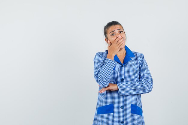 Junge Frau im blauen Gingham-Pyjamahemd, das Mund mit Händen bedeckt und überrascht, Vorderansicht schaut.