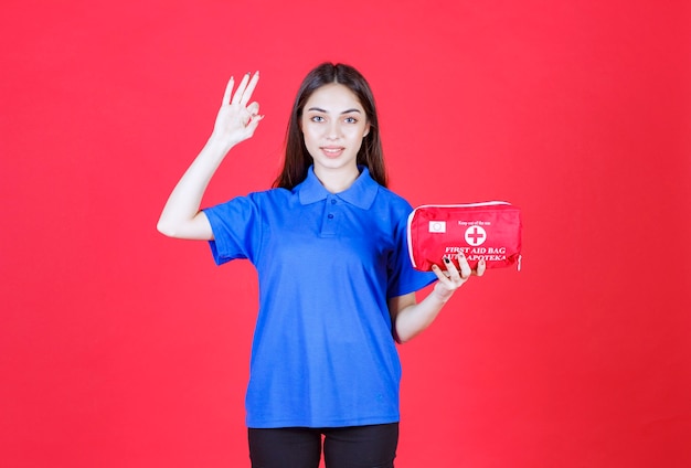 Junge Frau im blauen Hemd, die ein rotes Erste-Hilfe-Set hält und ein positives Handzeichen zeigt