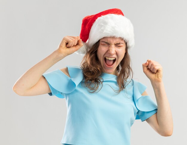 Junge Frau im blauen Oberteil und im Weihnachtsmannhut, die Fäuste ballen, die mit aggressivem Ausdruck verrückt verrückt schreien