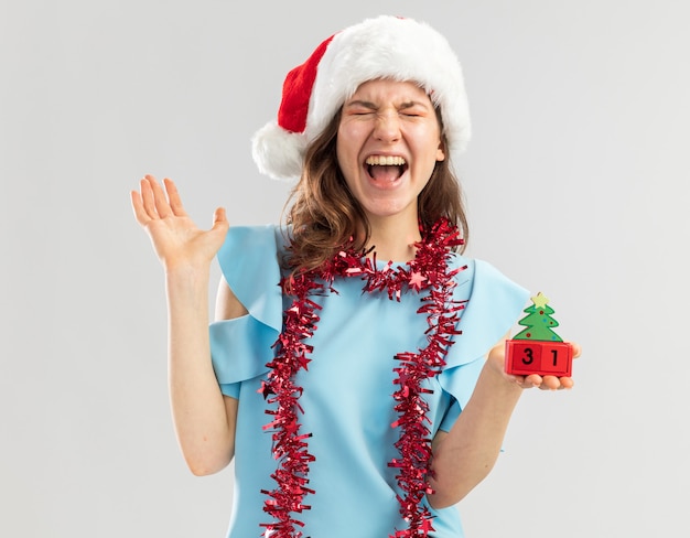 Junge Frau im blauen Oberteil und im Weihnachtsmannhut mit Lametta um ihren Hals, der Spielzeugwürfel mit glücklichem ney Jahresdatum hält, das verrückt glücklich schreit