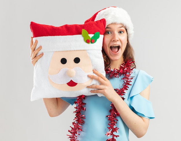 Junge Frau im blauen Oberteil und im Weihnachtsmannhut mit Lametta um ihren Hals, der Weihnachtskissen hält, das fröhlich und positiv lächelnd fröhlich schaut