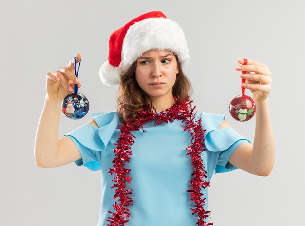 Junge Frau im blauen Oberteil und im Weihnachtsmannhut mit Lametta um ihren Hals, der Weihnachtskugeln hält, die verwirrt versuchen, Wahl zu treffen