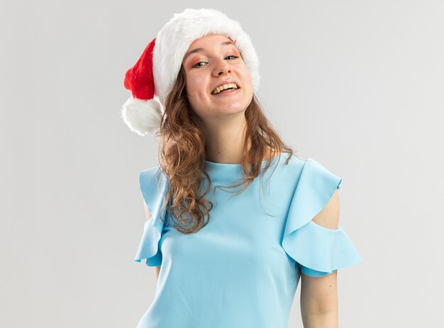 Junge Frau im blauen Oberteil und in der Weihnachtsmannmütze, die glücklich und fröhlich lächelnd zuversichtlich sieht