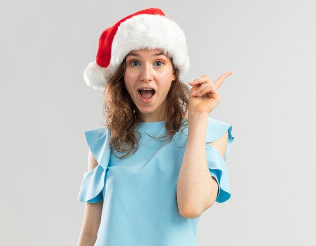 Junge Frau im blauen Oberteil und in der Weihnachtsmannmütze, die überrascht mit Lächeln auf klugem Gesicht sehen, das neue große Idee zeigt Zeigefinger zeigt
