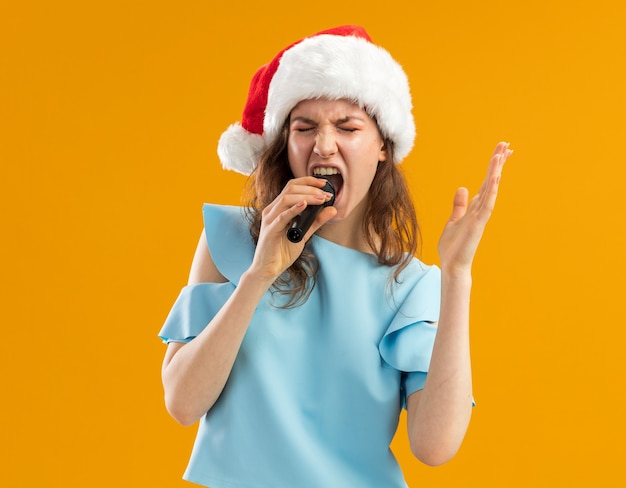 Junge Frau im blauen Oberteil und in der Weihnachtsmütze, die zum Mikrofon schreit, erregte verrückte emotionale mit erhobenem Arm