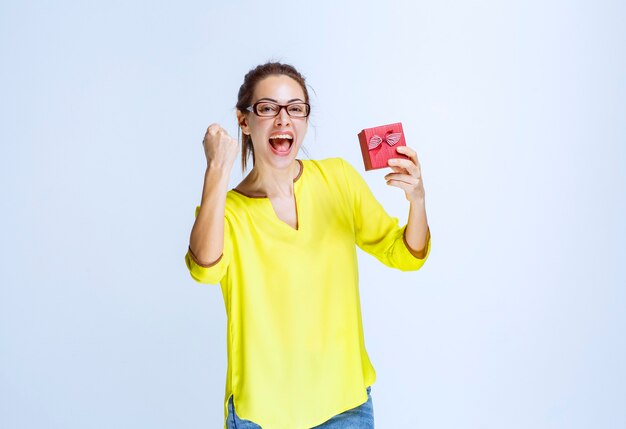 Junge Frau im gelben Hemd, die eine rote Geschenkbox hält und Freudenhandzeichen zeigt