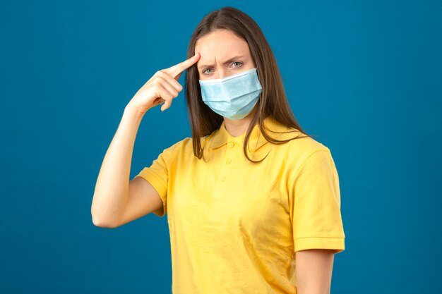 Junge Frau im gelben Poloshirt und in der medizinischen Schutzmaske, die mit dem Finger auf ihren missfallenen Blick des Kopfes auf lokalisiertem blauem Hintergrund zeigen