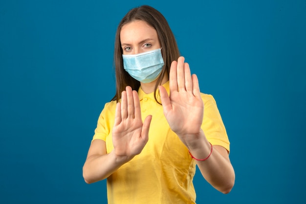 Junge Frau im gelben Poloshirt und in der medizinischen Schutzmaske, die Stoppgeste mit Händen auf lokalisiertem blauem Hintergrund machen