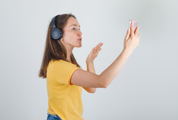 Junge Frau im gelben T-Shirt, Shorts, die Kuss am Telefon per Videoanruf senden