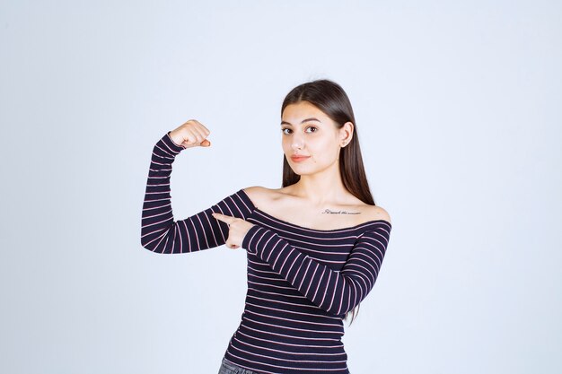 Junge Frau im gestreiften Hemd, das ihren Armmuskel und ihre Faust zeigt