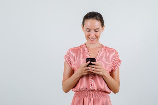 Junge Frau im gestreiften Kleid, das auf Handy schreibt und froh schaut, Vorderansicht.