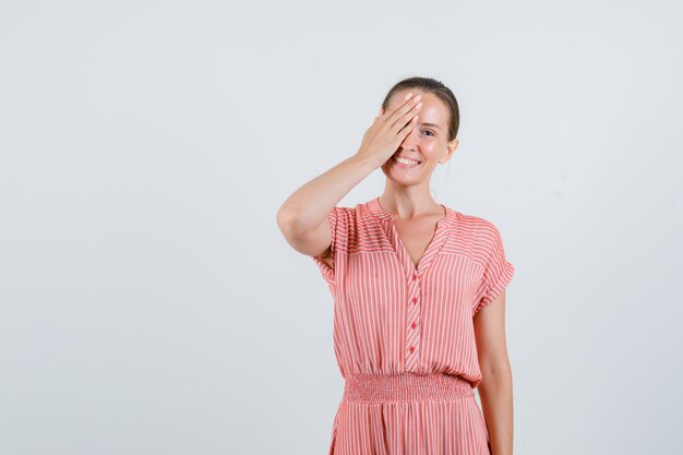 Junge Frau im gestreiften Kleid, das Auge mit Hand bedeckt und fröhlich, Vorderansicht schaut.