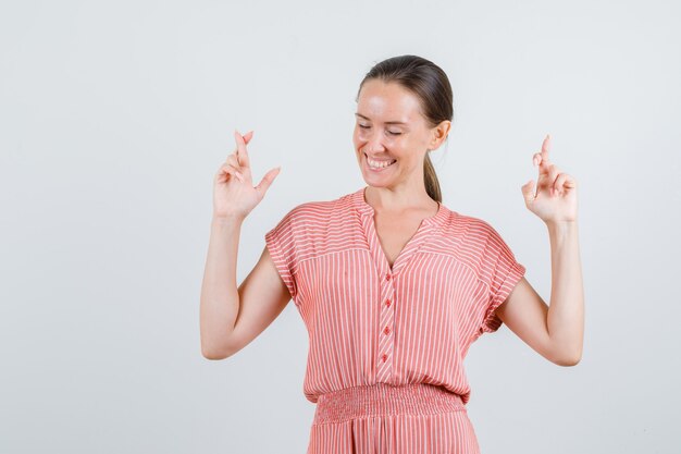 Junge Frau im gestreiften Kleid, die Finger gekreuzt hält und fröhlich, Vorderansicht schaut.