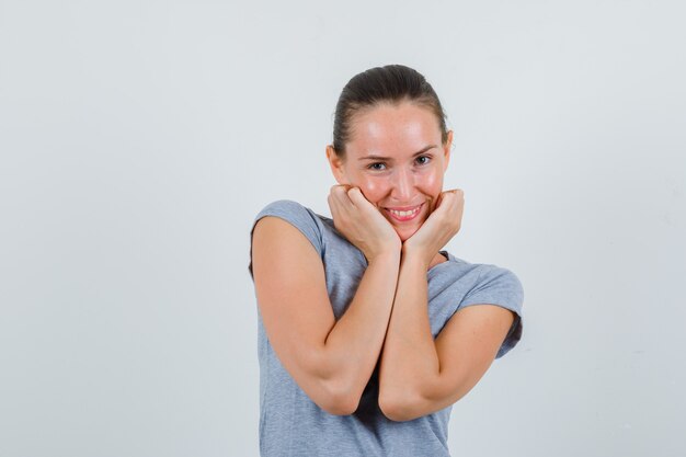 Junge Frau im grauen T-Shirt, das Kinn auf Handflächen stützt und niedliche Vorderansicht schaut.