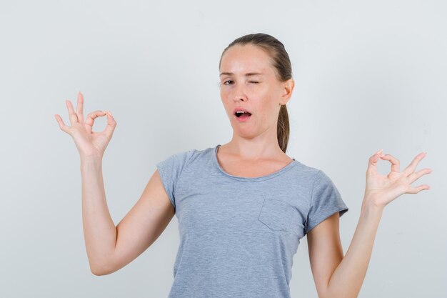 Junge Frau im grauen T-Shirt zeigt ok Geste und zwinkerndes Auge, Vorderansicht.