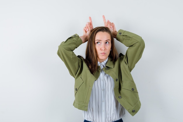 Kostenloses Foto junge frau im hemd, jacke, die die finger als stierhörner über dem kopf hält und nachdenklich aussieht, vorderansicht.