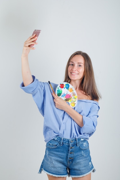 Junge Frau im Hemd, Shorts, die Selfie mit Malwerkzeugen nehmen