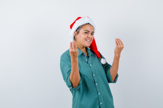 Junge Frau im Hemd, Weihnachtsmütze, die italienische Geste zeigt und fröhlich aussieht, Vorderansicht.