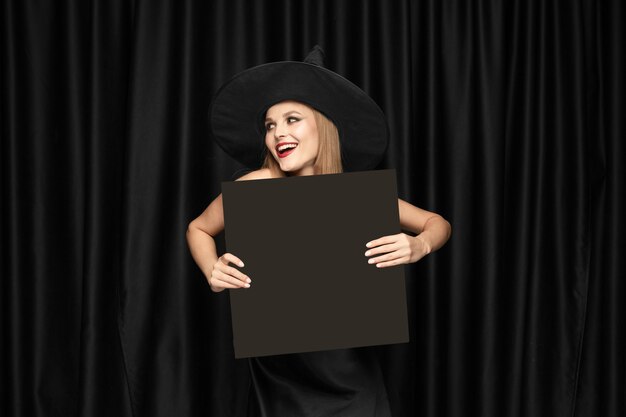 Junge Frau im Hut als Hexe, die schwarze Tafel gegen schwarze Vorhänge hält