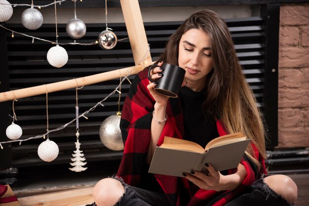 Junge Frau im karierten Plaid, die auf dem Boden sitzt und ein Buch liest.