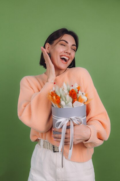 Junge Frau im lässigen Pfirsichpullover lokalisiert auf grüner Olivenwand halten orange weiße Blumenkastenzusammensetzung von Baumwollblumen Gypsophila Weizen und Lagurus für ein Geschenk glücklich erstaunt überrascht