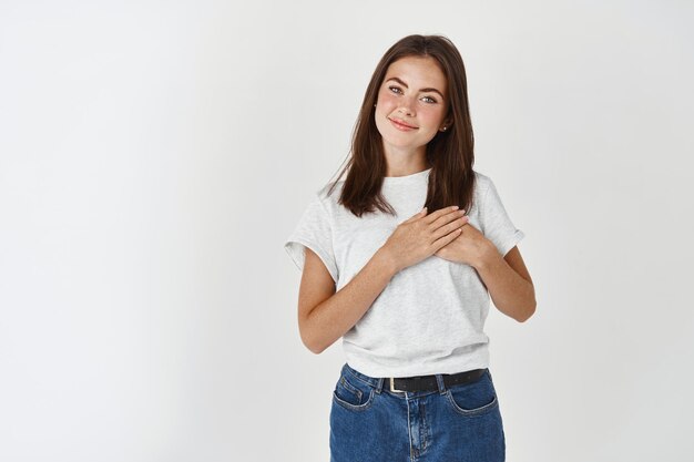 Junge Frau im lässigen T-Shirt, die Hände auf dem Herzen hält und mit Sorgfalt und Liebe lächelt und über weißer Wand steht