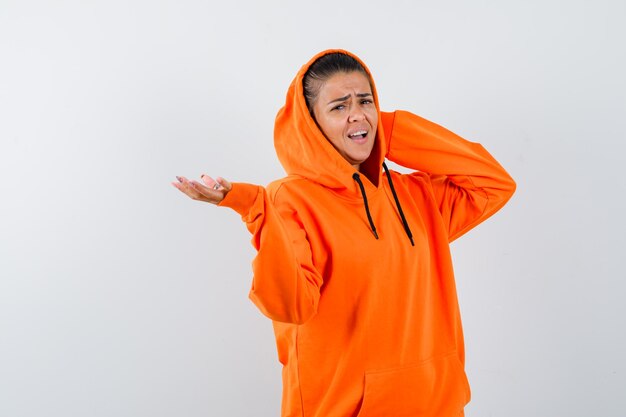 Junge Frau im orangefarbenen Hoodie, die eine Hand streckt, als sie etwas hält und die Hand hinter dem Hals hält und gehetzt aussieht