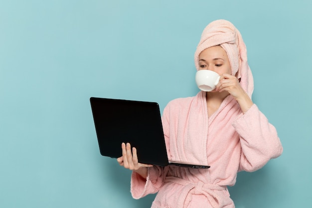 junge Frau im rosa Bademantel nach der Dusche, die Kaffee trinkt und Laptop auf Blau verwendet