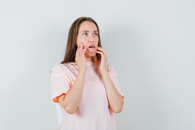 Junge Frau im rosa T-Shirt, das Finger auf Wangen hält und nachdenklich, Vorderansicht schaut.