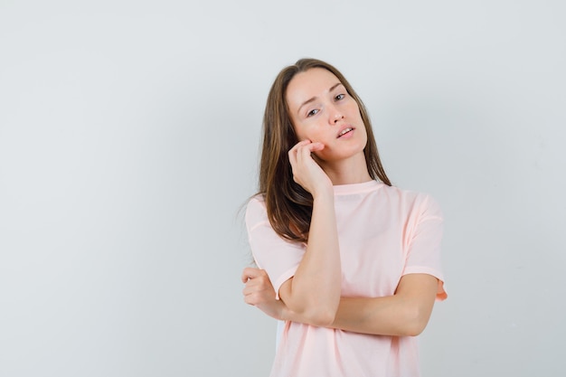 Junge Frau im rosa T-Shirt, das ihre Gesichtshaut auf Wange berührt und faszinierend, Vorderansicht schaut.