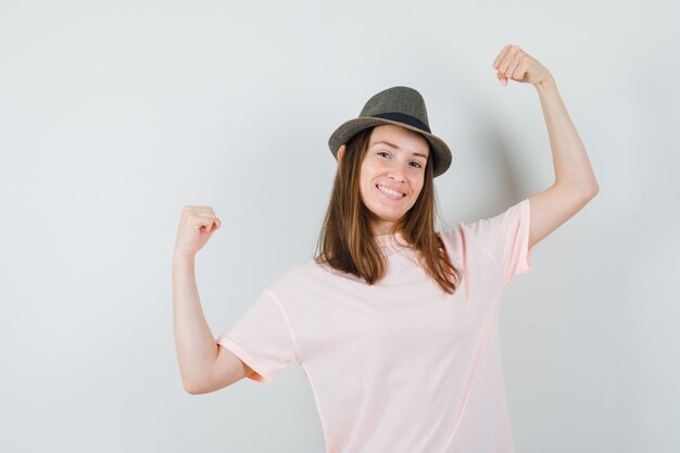 Junge frau im rosa t-shirt, hut, der siegergeste zeigt und glückselig schaut, vorderansicht.