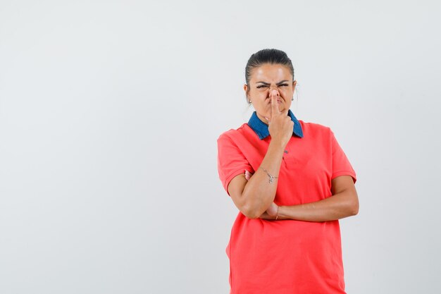 Junge Frau im roten T-Shirt, das in der denkenden Haltung steht und Zeigefinger auf Nase setzt und gehetzt, Vorderansicht schaut.