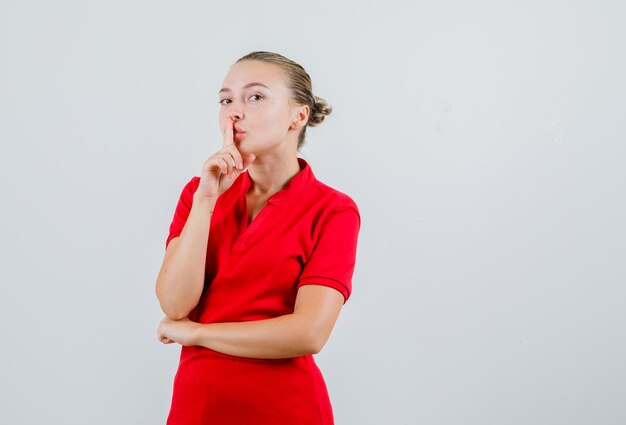 Junge Frau im roten T-Shirt, das Schweigengeste zeigt und vorsichtig schaut