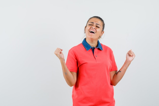 Junge Frau im roten T-Shirt, das Siegergeste zeigt und glückliche Vorderansicht schaut.