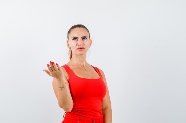 Junge Frau im roten Trägershirt, Hosen, die Hand in fragender Geste strecken und ernst schauen, Vorderansicht.