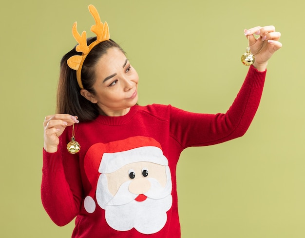 Junge Frau im roten Weihnachtspullover, der lustigen Rand mit Hirschhörnern hält, die Weihnachtskugeln halten, die verwirrt suchen, Zweifel haben