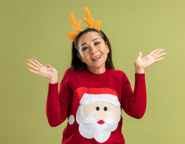 Junge Frau im roten Weihnachtspullover, der lustigen Rand mit Hirschhörnern trägt, die mit glücklichem Gesicht schauen, das mit erhobenen Armen lächelt