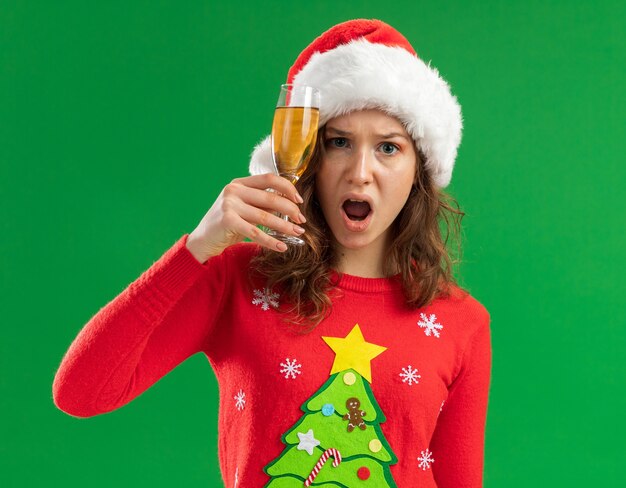 Kostenloses Foto junge frau im roten weihnachtspullover und in der weihnachtsmannmütze, die glas champagner halten, betrachten kamera genervt und irritiert, die über grünem hintergrund stehen